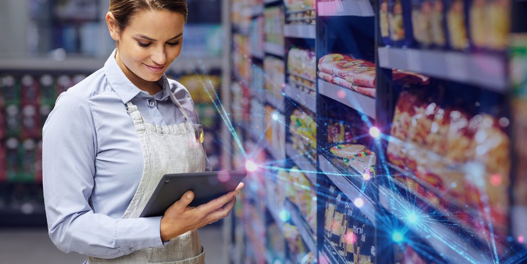 A retail employee holding a tablet while analyzing product shelves in a grocery store, representing data-led field marketing and compliance.