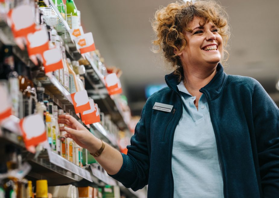A smiling retail employee in a store, interacting with shelves of FMCG products, showcasing in-store compliance and visibility.