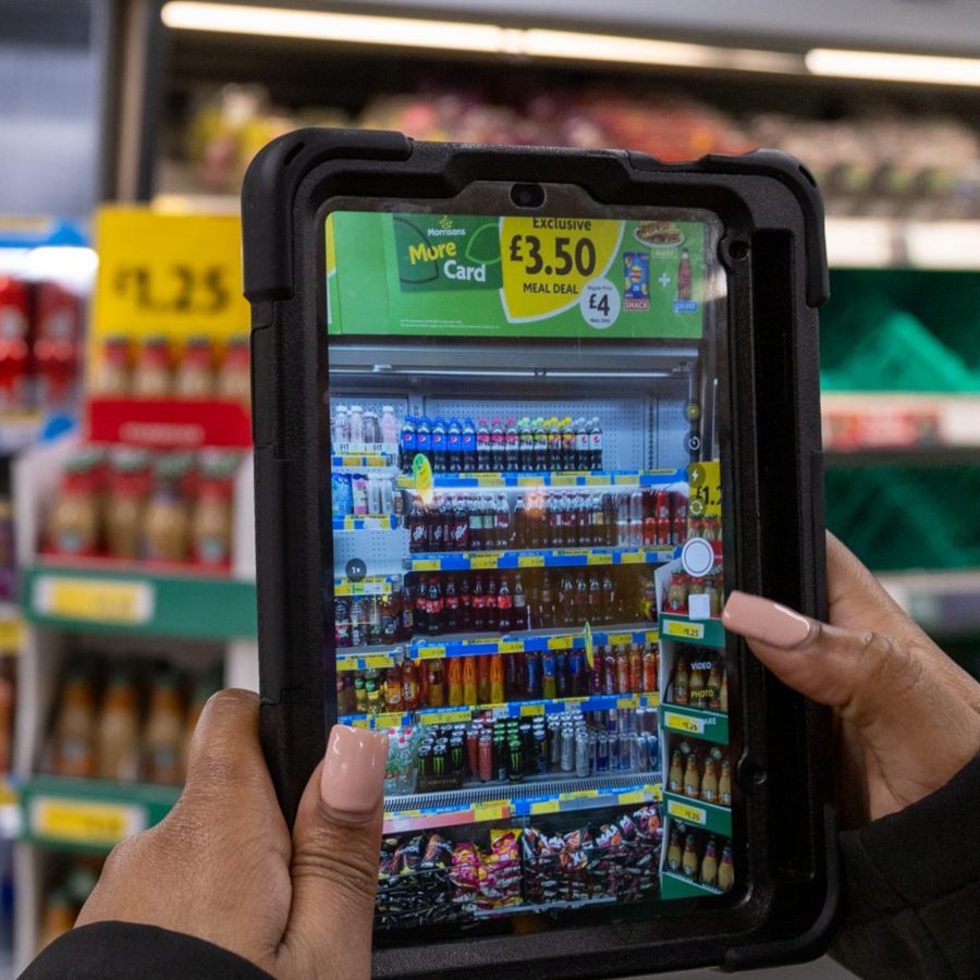 A person holding a tablet displaying an organized shelf of beverages in a grocery store, highlighting promotional deals and product visibility for FMCG brands.