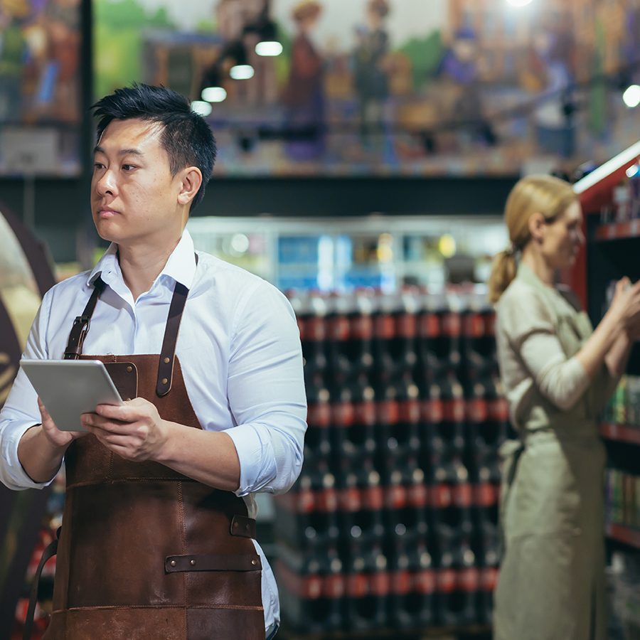 A male retail employee in an apron holding a tablet, overseeing product placement in a grocery store, showcasing effective in-store compliance and promotional strategies.